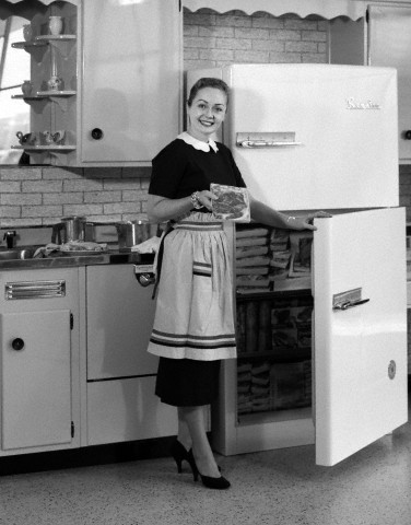 1950s smiling woman housewife in kitchen taking frozen food out of refrigerator freezer.