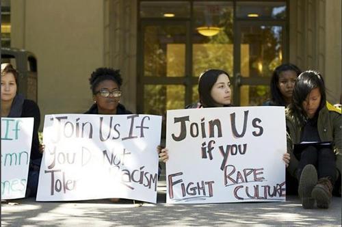 Photo of a row of young women sitting outside, one holding a sign that says 'Join us if you fight rape culture'