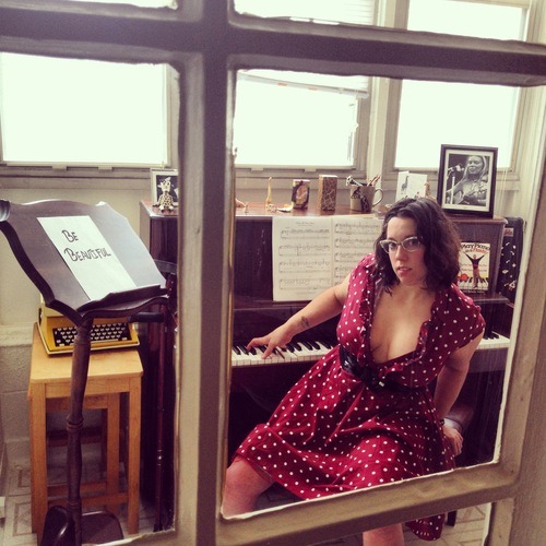 Natalie is in the sunroom of her apartment, sitting sideways on a piano bench. Her right hand is playing an upright 1930s Hamilton piano, which is directly behind her. Her left hand is positioned behind her for balance. To the left of the piano is a wooden music stand with a white piece of paper on it that says “Be Beautiful.” Behind the music stand is a vintage yellow typewriter. The sheet music is Tori Amos’ “Silent All These Years.” Natalie identifies strongly with the lyrics. On top of the piano is her collection of giraffes and a framed black and white photo of Joni Mitchell, her childhood muse. Natalie is looking sideways at the camera, wearing a burgundy and white polka dot dress, which is unbuttoned to show her cleavage. She is a white woman with dark shoulder-length hair and glasses. Denise is taking the photo through the glass door of the sunroom, so that Natalie’s body is partially framed by the white panels of the door. One crutch is leaning up against the piano. Natalie’s crutches are also burgundy.