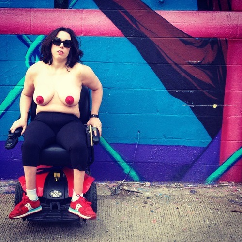 This photo was taken outside of The Fridge DC, a local art gallery and performance venue. Natalie is posing in front of a mural in her electric wheelchair. The mural is a series of barbed wire, pipes and trees in blues, purples and turquoise. The photo is cropped so that the wheelchair is positioned to the far left of the mural. Natalie is wearing red sneakers, black leggings, red pasties over her nipples, and Warby Parker sunglasses. Natalie’s hands are holding onto the arms of the wheelchair and pushing her shoulders back. Her feet are turned outward at an angle that is consistent with Cerebral Palsy. Denise asked Natalie to give her a “bitch don’t play face” – so she looks tough.