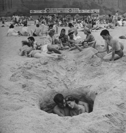  Young couple cuddling as they sit down in a hole in the sand while others lie around behind them on a hot Independence Day at the beach, 1949. 